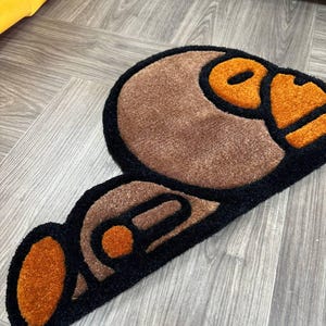 May include: A brown and black rug with a cartoon-like design of a monkey face. The monkey's face is outlined in black and has orange eyes and a mouth.