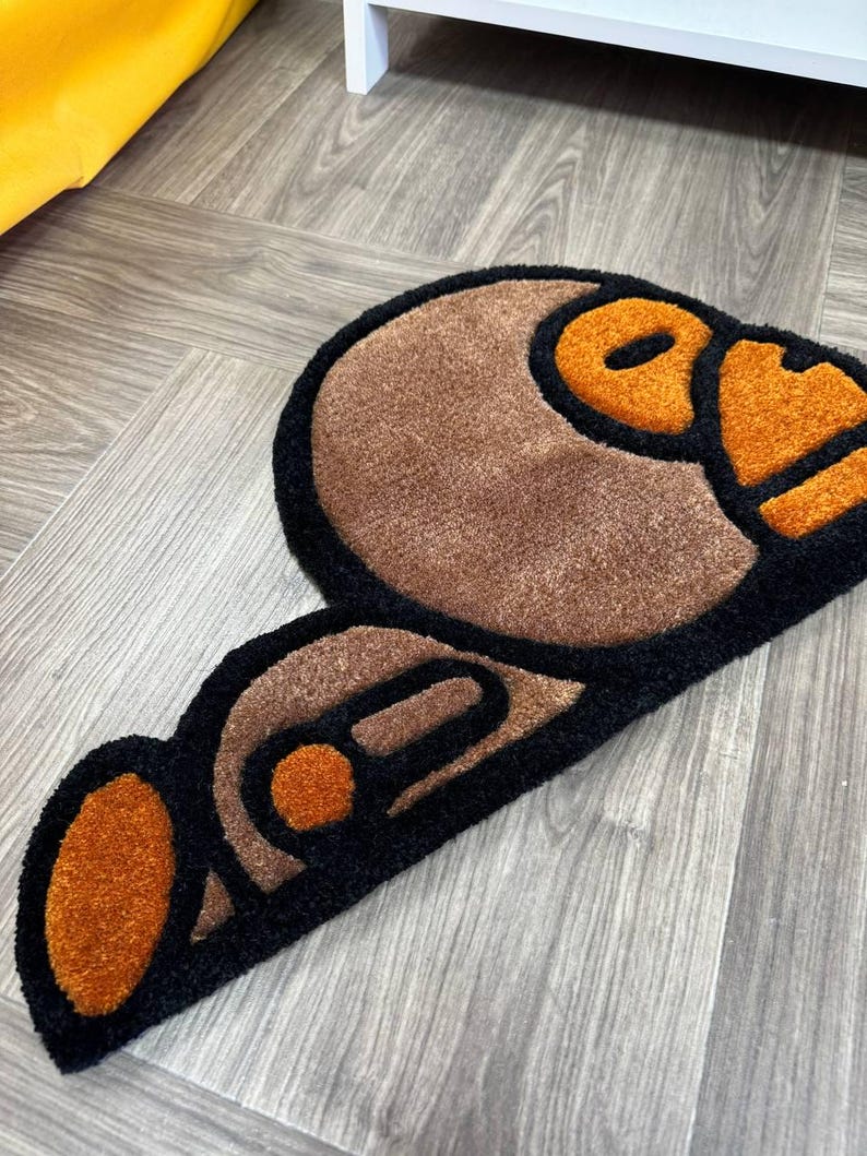 May include: A brown and black rug with a cartoon-like design of a monkey face. The monkey's face is outlined in black and has orange eyes and a mouth.