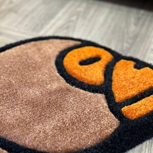 May include: A brown and black rug with a large orange and black design. The design is a stylized cartoon face with a large orange eye.