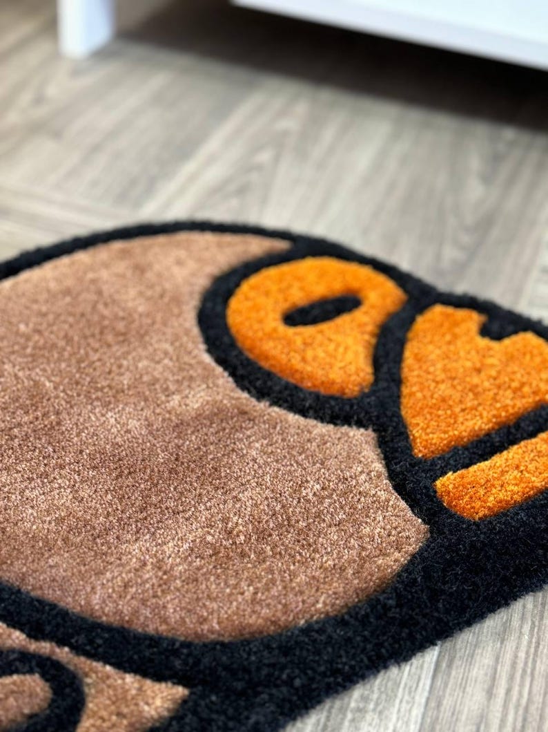 May include: A brown and black rug with a large orange and black design. The design is a stylized cartoon face with a large orange eye.