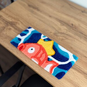 May include: A rectangular, colorful rug featuring a cartoon fish design. The rug has a blue background with white and light blue wave patterns. The fish is red with white accents and a yellow star. The rug is placed on a wooden surface.