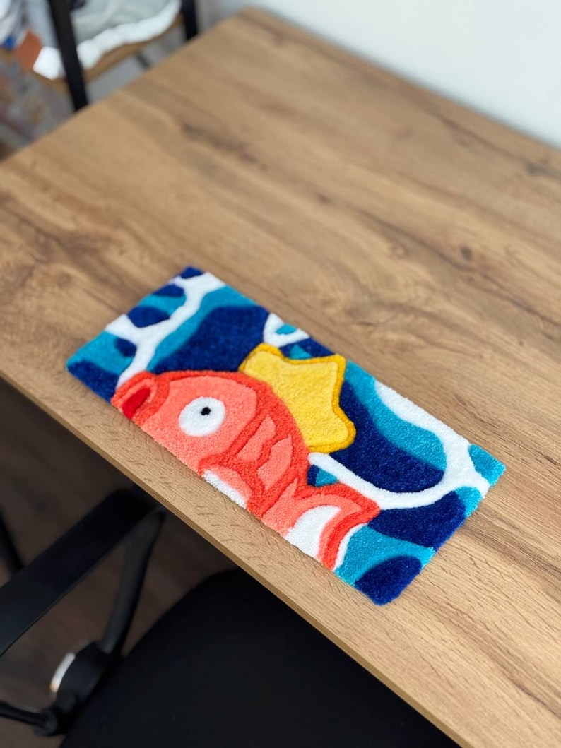 May include: A rectangular, colorful rug featuring a cartoon fish design. The rug has a blue background with white and light blue wave patterns. The fish is red with white accents and a yellow star. The rug is placed on a wooden surface.