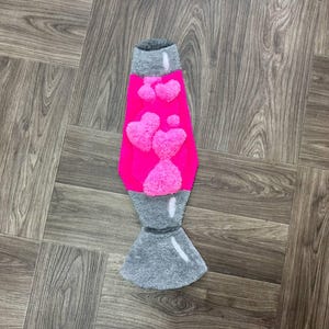 May include: A pink lava lamp rug with a gray base and white heart-shaped designs. The rug is made of fluffy material and is shaped like a lava lamp.