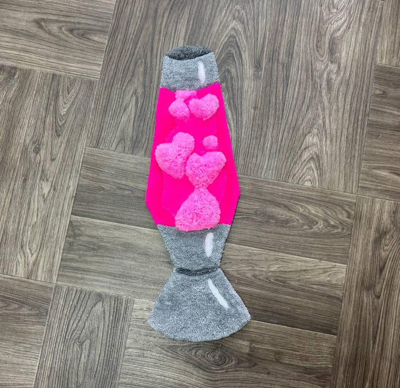 May include: A pink lava lamp rug with a gray base and white heart-shaped designs. The rug is made of fluffy material and is shaped like a lava lamp.