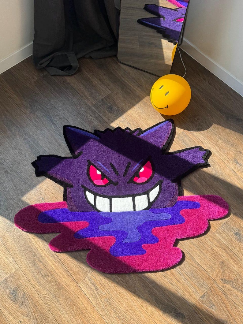 Puede incluir: Una alfombra de mechones con forma personalizada que presenta al personaje morado Gengar con orejas puntiagudas, ojos rojos brillantes y una amplia sonrisa blanca. El personaje parece derretirse en un charco vibrante de tonos morado y magenta.