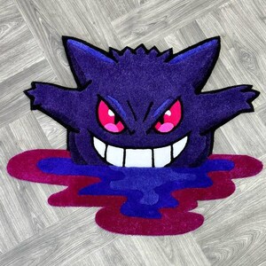 Puede incluir: Una alfombra tufting con la forma del personaje Gengar, que presenta un cuerpo de color morado intenso, orejas puntiagudas y una amplia sonrisa con dientes blancos. El personaje tiene ojos de color rosa brillante y emerge de un charco de l&iacute;quido azul oscuro y magenta.