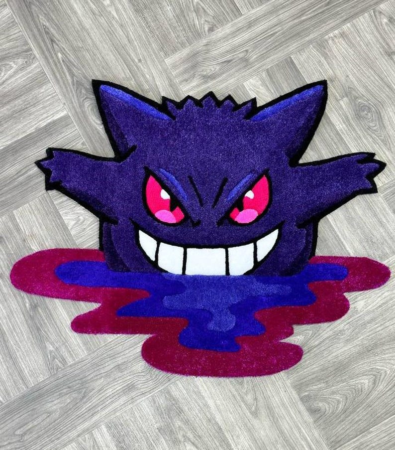 Puede incluir: Una alfombra tufting con la forma del personaje Gengar, que presenta un cuerpo de color morado intenso, orejas puntiagudas y una amplia sonrisa con dientes blancos. El personaje tiene ojos de color rosa brillante y emerge de un charco de l&iacute;quido azul oscuro y magenta.