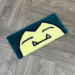 Puede incluir: Una alfombra amarilla y verde con una cara de dibujos animados de un personaje Pokemon. El personaje tiene una amplia sonrisa con dientes blancos y los ojos cerrados.