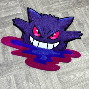 Puede incluir: Una alfombra tufted con la forma del personaje Gengar, que presenta un cuerpo de color morado intenso, orejas puntiagudas y una amplia sonrisa con dientes blancos. El personaje tiene ojos de color rosa brillante y parece emerger de un charco de l&iacute;quido morado y magenta.