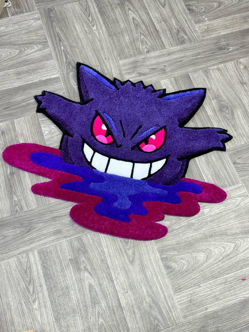 Puede incluir: Una alfombra tufted con la forma del personaje Gengar, que presenta un cuerpo de color morado intenso, orejas puntiagudas y una amplia sonrisa con dientes blancos. El personaje tiene ojos de color rosa brillante y parece emerger de un charco de l&iacute;quido morado y magenta.