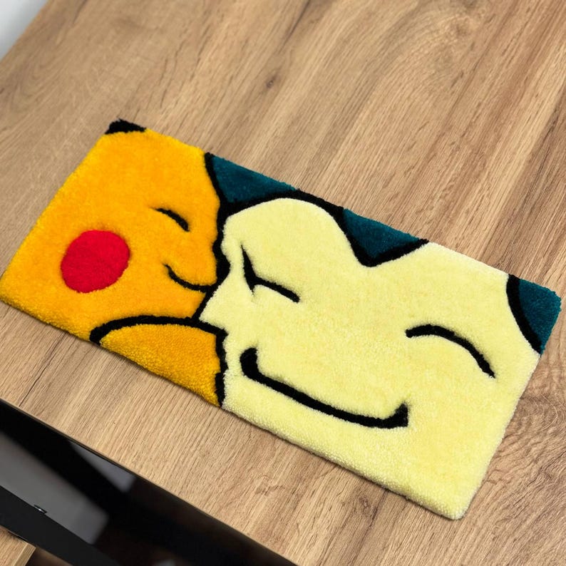 Może przedstawiać: Prostokątny dywan z ż&oacute;łtym wzorem Pikachu z czerwonym policzkiem i uśmiechniętą twarzą, obok jasnoż&oacute;łtej postaci z uśmiechniętą twarzą. Dywan ma ciemnozieloną obw&oacute;dkę i jest umieszczony na drewnianej powierzchni.