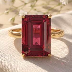 Puede incluir: Un anillo de oro con una gran piedra preciosa de color rojo intenso, de corte esmeralda rectangular y sujeta por cuatro garras. La banda es lisa y pulida, con un diseño sencillo que resalta el color vibrante y la claridad facetada.