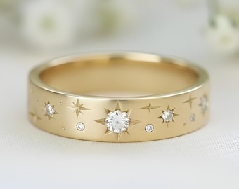 Op de afbeelding: Een gouden bandring met een hemels ontwerp. De ring is versierd met verschillende stervormen en kleine, ronde diamanten. De centrale ster is groter en prominenter. De ring is geplaatst tegen een zachte, vage achtergrond van witte bloemen.