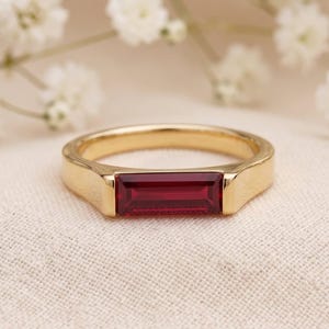 Peut inclure: Une bague en or ornée d'une pierre précieuse rouge foncé taille baguette disposée horizontalement au centre. L'anneau est lisse et poli, présentant une finition métallique nette qui complète la pierre rectangulaire éclatante. Le design est simple.
