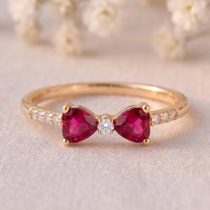 Op de afbeelding: Een gouden ring met een centraal strikontwerp bestaande uit twee hartvormige rode edelstenen en een kleine ronde witte edelsteen in het midden. De ringband is aan weerszijden van de strik versierd met een reeks kleine, heldere ronde edelstenen.