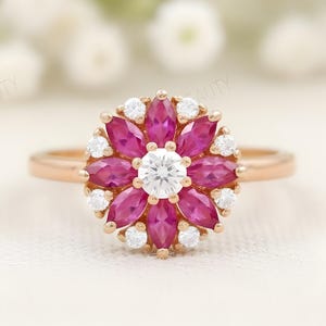 May include: A rose gold ring featuring a flower-shaped design. The center stone is a round, white diamond, surrounded by marquise-cut, pink gemstones and smaller white diamonds. The ring is set against a soft, white background.