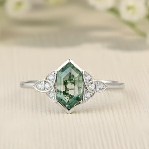 May include: A silver ring with a hexagonal green gemstone and white detailing. The ring has a slender band, with small white stones flanking the central gem. The ring is set against a soft, neutral background with white flowers.