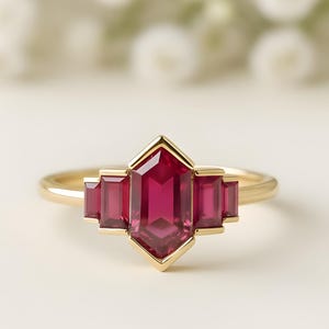 May include: A gold ring with a central, faceted, ruby-red gemstone in a geometric design. The ring has a central, elongated stone flanked by two smaller stones on each side. The background is a soft, blurred image of white flowers.