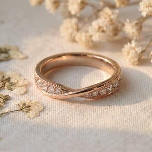 Peut inclure: Une bague en or rose présentant un design de bande torsadée avec une série de petites pierres claires taillées en rond serties sur le devant. Le métal a une finition polie et l'anneau comprend des détails milgrain délicats sur les bords.