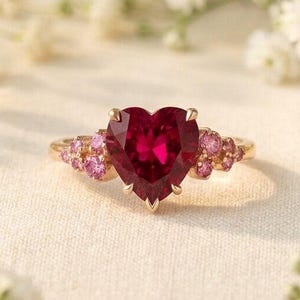 May include: A gold ring featuring a large, heart-shaped, deep red gemstone at the center, flanked by smaller, round, pink accent stones on each side. The ring is set on a thin, polished gold band, showcasing a delicate and symmetrical design.