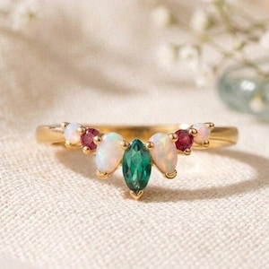 May include: A gold ring featuring a central marquise-cut green gemstone flanked by two pear-cut opals and two round-cut red rubies. The stones are set in a curved arrangement along the top of the thin, polished gold band, creating a symmetrical design.