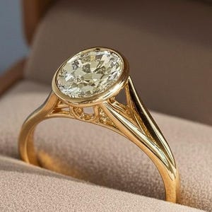 May include: A gold ring featuring an oval-cut diamond in a bezel setting. The ring is presented in a brown box with a beige interior. The band has a split shank design.