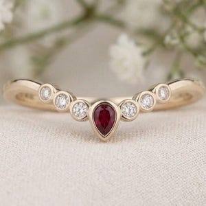 Puede incluir: Un anillo de oro curvado con un rubí rojo central en forma de pera, flanqueado por tres diamantes blancos redondos a cada lado. Las piedras están engastadas en bisel a lo largo de la banda, creando un diseño de arco simétrico y pulido.