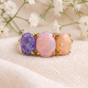 May include: A gold ring featuring three oval-cut stones set in a row. The left stone is a vibrant purple with iridescent flecks, the center stone is a soft pastel pink, and the right stone is a warm peach color with subtle light reflections.