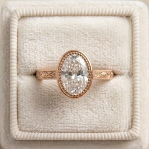 May include: Rose gold engagement ring with an oval-cut diamond. The ring features a beaded bezel setting and a detailed band with an engraved pattern. The ring is presented in a white velvet ring box.