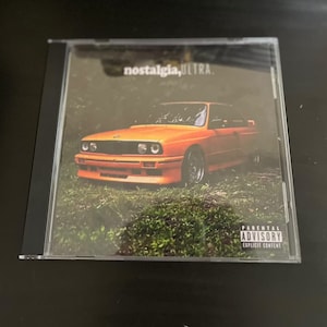 May include: A CD case with a bright orange car on the front. The car is in a grassy field. The text "nostalgia, ULTRA." is printed on the case. A parental advisory sticker is on the bottom right corner.