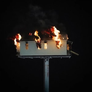 May include: A small, light-colored house on a tall pole is engulfed in flames against a black background. The fire is bright orange and yellow, with smoke rising above the burning structure. The house has several windows and a simple design.