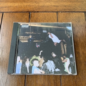 May include: A CD case with a black spine and a clear front cover. The cover art features a group of people in a dimly lit room. One person is hanging upside down from the ceiling. The text "Parental Advisory Explicit Content" is printed in the upper left corner of the cover.