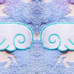 Könnte beinhalten: Zwei weiße Filzflügel mit blauen gestickten Kanten auf einem blauen und violetten flauschigen Hintergrund.