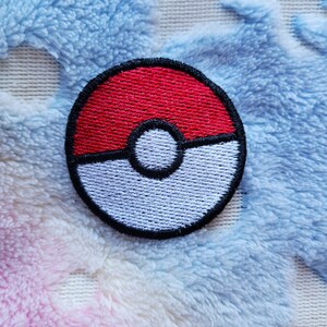 Puede incluir: Un parche bordado de una Poké Ball, un objeto circular con la parte superior roja y la inferior blanca con un círculo negro en el centro.