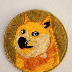 Könnte beinhalten: Ein runder gestickter Aufnäher mit einem Shiba Inu Hund mit einem überraschten Ausdruck. Der Hund ist gelb und orange mit schwarzen Augen und einer schwarzen Nase. Der Hintergrund des Aufnähers ist dunkelgrün.