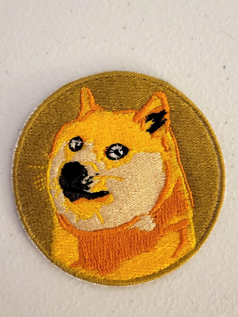 Puede incluir: Un parche redondo bordado con un perro Shiba Inu con una expresi&oacute;n de sorpresa. El perro es amarillo y naranja con ojos negros y una nariz negra. El fondo del parche es verde oscuro.