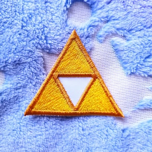 Könnte beinhalten: Ein goldfarbener gestickter Dreiecks-Patch mit einem weißen inneren Dreieck. Der Patch befindet sich auf einem blauen und weißen flauschigen Stoff.