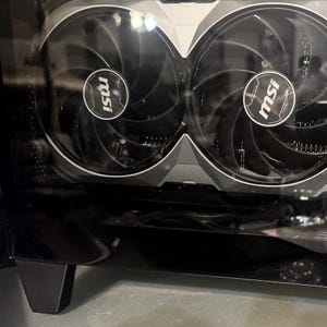 Puede incluir: Primer plano de un componente de computadora negro con dos ventiladores grandes. Cada ventilador tiene un centro con el logotipo "msi" en blanco. Los ventiladores están rodeados por un marco plateado y negro, y el componente está alojado en una carcasa negra.