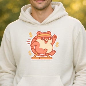 May include: Cream-colored hoodie featuring a cartoon hamster wearing sunglasses and a headband, running on a hamster wheel. The design includes dollar signs and speed lines, creating a playful and energetic aesthetic. A fun, casual apparel item.