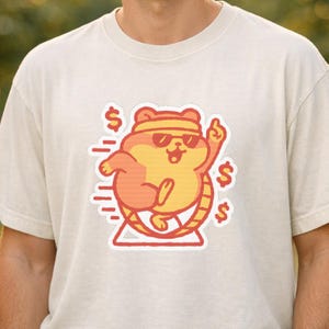 May include: A beige t-shirt featuring a cartoon hamster in a hamster wheel, wearing sunglasses and a headband. The hamster is giving a thumbs up, with dollar signs around it. The design is in shades of orange and yellow.