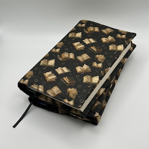 May include: A fabric book cover featuring a repeating pattern of open books in shades of gold and tan against a black background. The cover is wrapped around a closed book, with a thin black ribbon bookmark extending from the bottom edge.