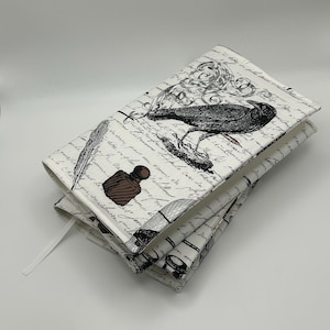 May include: A white book cover with a black crow illustration, inkwell, and script design. The cover has a white ribbon bookmark. The design is repeated on the folded cover. The book cover is made of fabric.