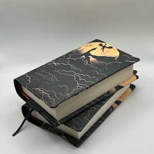 May include: A stack of three black hardback books with gold and white illustrations of clouds and a dragon flying in front of a yellow moon. The top book has a black ribbon bookmark.