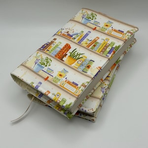 May include: A fabric book cover featuring a repeating pattern of horizontal shelves filled with colorful books, potted plants, and small ceramic teacups. The design includes various shades of green, orange, blue, and yellow on a cream background.