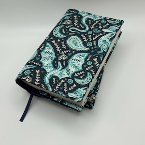 Peut inclure: Une couverture de livre en tissu avec un fond bleu marine foncé orné d'un motif répétitif de dragons, de croissants de lune, de formes cachemire et de petites tiges florales en bleu sarcelle et blanc. Un marque-page en ruban bleu marine dépasse.