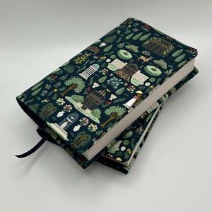 May include: Two hardcover books with a dark teal cover featuring a garden design with trees, birdcages, and architectural elements. The books have white pages and a dark blue ribbon bookmark. The covers have a repeating pattern of green and white elements.