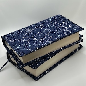 May include: Two hardback books with navy blue covers displaying a white constellation design. The covers have a fabric texture and a matching navy blue bookmark. The books are stacked one on top of the other.