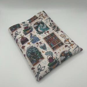 May include: A rectangular fabric sleeve featuring a cream background with a repeating pattern of illustrations including a bookshelf, a dragon, potion bottles, scissors, mushrooms, and a teapot. The sleeve has a single white snap button closure.