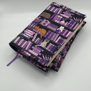 May include: A fabric book cover featuring a repeating pattern of bookshelves filled with purple and black books, various cats, and text including "9 LIVES," "BIG CATS," "FISHING," and "MISCHIEF." The cover includes a purple ribbon bookmark.