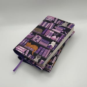 May include: A book cover with a black background featuring a bookshelf design with purple and white books, and various cat illustrations. The cover has a purple bookmark. The book is closed.